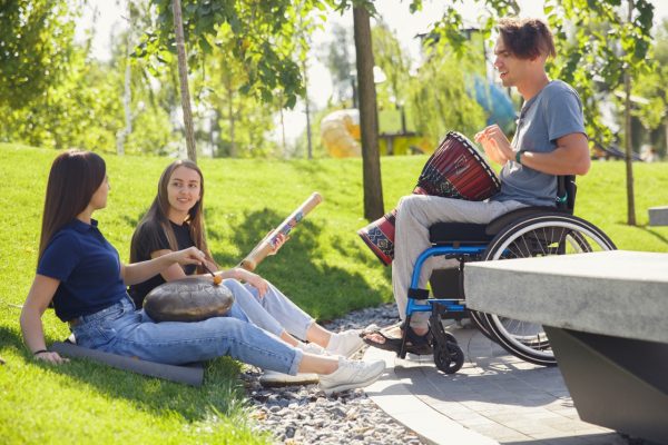 Happy handicapped man on a wheelchair spending time with friends playing live instrumental music outdoors. Concept of social life, friendship, possibilities, inclusion, diversity.