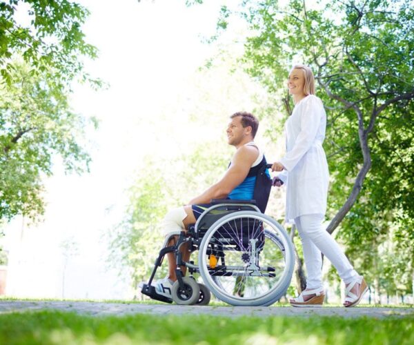 Pretty nurse walking with male patient in a wheelchair in park